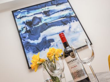 A bottle of red wine with two wine glasses and yellow flowers in a vase in front of a blue abstract painting on a wall