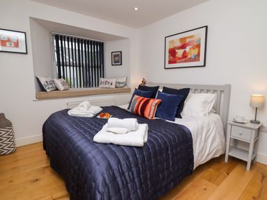 A bedroom with a bed with pillows and folded towels a window seat with cushions and framed pictures on white walls at The View in St Ives