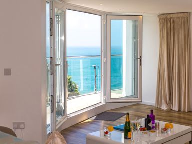 A room with an open glass door leading to a balcony overlooking the ocean with a table holding drinks and snacks at Portside in Carbis Bay