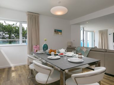 A dining table set with plates cups and croissants in a room with white chairs a sofa and large windows at Portside in Carbis Bay
