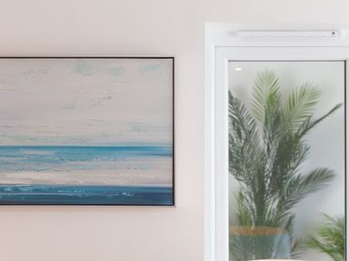 A framed abstract painting with blue and beige tones on a wall next to a white framed glass door with a green plant visible through the glass at Portside in Carbis Bay