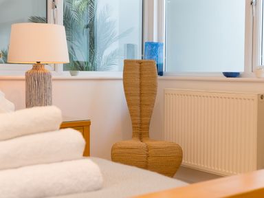A corner of a room with a woven chair a table lamp and a stack of towels at Portside in Carbis Bay