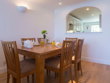 A dining room with a wooden table and six wooden chairs with beige cushions next to a kitchen with white cabinets at Sapphire in Carbis Bay
