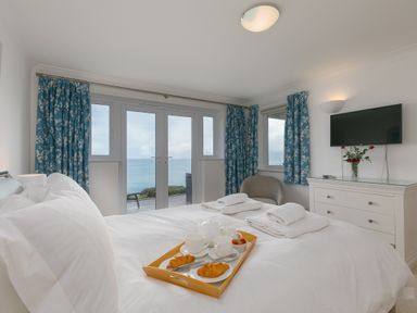 A bedroom with a bed and breakfast tray with croissants and cups by windows with blue floral curtains and a dresser with flowers at Sapphire in Carbis Bay
