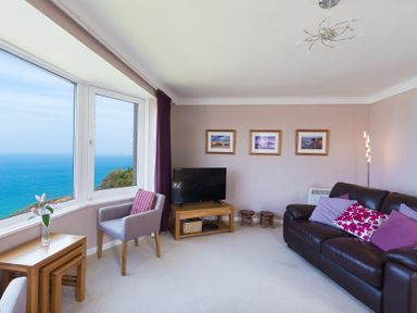 A living room with a large window overlooking the ocean a leather sofa with cushions a TV on a wooden stand a chair and nested wooden tables at Seathrift in Carbis Bay