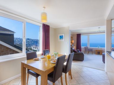 A dining table set with croissants and juice near a window with a view of houses and a living room with chairs and a sea view at Seathrift in Carbis Bay