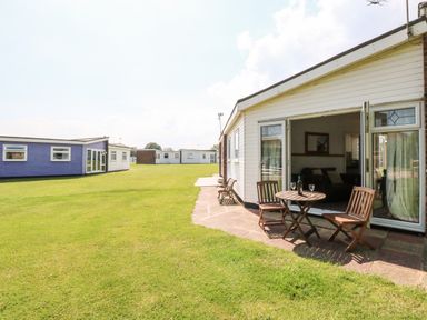 Exterior view of a white chalet with an open door leading to a patio with a wooden table and two chairs on a lawn at Chalet 150 in Winterton-On-Sea