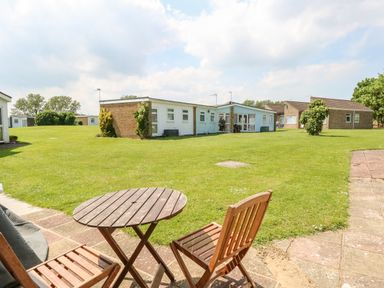 A patio with wooden table and chairs overlooking a grassy area and low buildings at Chalet 150 in Winterton-On-Sea