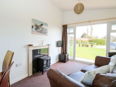 A living room with a brown leather sofa a small black fireplace a wall-mounted TV and large sliding glass doors at Chalet 150 in Winterton-On-Sea
