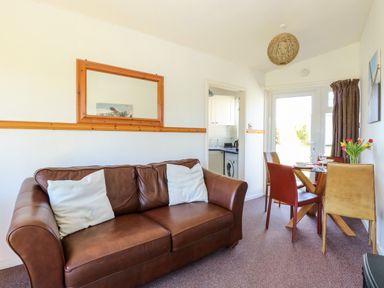 A living room with a brown leather sofa two white cushions a wooden framed mirror and a dining table with four chairs and flowers at Chalet 150 in Winterton-On-Sea