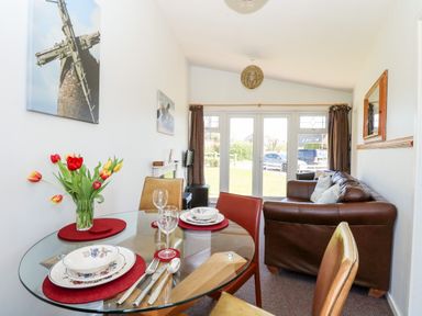 A dining area with a glass round table set with plates and glasses next to a living room with a brown leather sofa and glass doors at Chalet 150 in Winterton-On-Sea