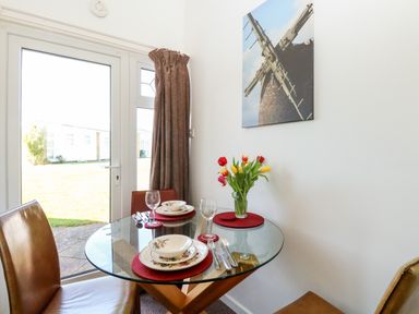 A dining area with a round glass table set with plates glasses and cutlery brown chairs a vase with flowers and a wall picture of a windmill at Chalet 150 in Winterton-On-Sea