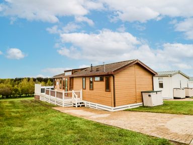 A wooden lodge with a white railing and steps on a paved pathway surrounded by grass at Woodland View Lodge Swarland near Felton