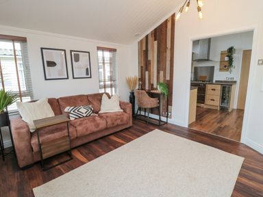 A living room with a brown sofa a small side table two framed pictures and a desk leading to a kitchen at Woodland View Lodge Swarland near Felton