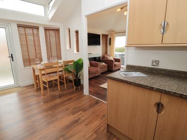 A dining area with a wooden table and chairs next to a living room with two armchairs and a wall-mounted TV at Woodland View Lodge in Swarland near Felton