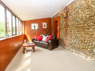 A room with wood panel walls stone wall a brown leather sofa with red pillows a wooden coffee table and large windows at The Hermitage in West Beckham near Sheringham