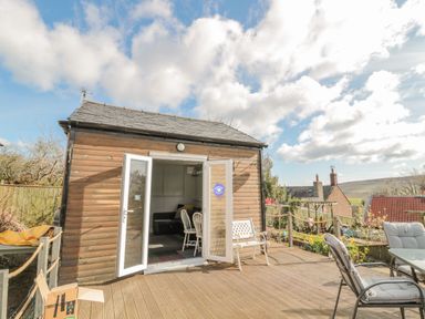 A wooden garden shed with open white double doors on a wooden deck with outdoor chairs and a bench at Woolley Lodge in Wooler