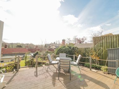 An outdoor wooden deck with four cushioned chairs around a glass table and a wooden fence with plants at Woolley Lodge in Wooler