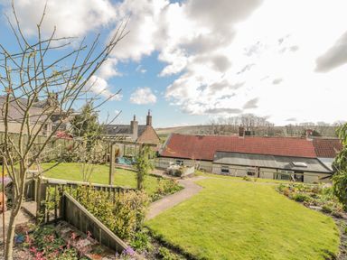 A garden with grass path wooden fence bushes and houses with tiled roofs in the background at Woolley Lodge in Wooler