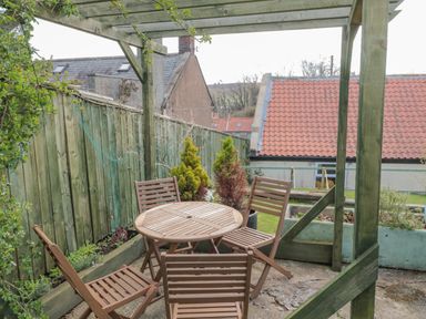 An outdoor patio with a round wooden table and four wooden chairs under a wooden pergola with plants and a wooden fence near Woolley Lodge in Wooler