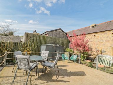 An outdoor patio with a glass table and four cushioned chairs two teal chairs pink flowering bush and a metal shed at Woolley Lodge in Wooler