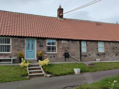 Exterior of a stone cottage with a red tiled roof green door steps and a bench at Woolley Lodge in Wooler