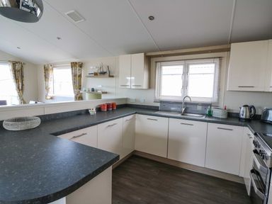 A kitchen with white cabinets black countertops a sink and window blinds with a dining area and windows with patterned curtains visible at The Potting Shed in New Quay