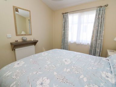 A bedroom with a bed covered in floral bedding a window with floral curtains a wall mirror and a small shelf at The Potting Shed in New Quay