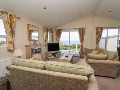A living room with two sofas a coffee table a TV on a stand a fireplace and yellow floral curtains with a view outside at The Potting Shed in New Quay