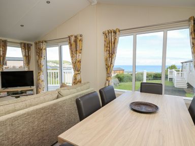 A dining table with chairs and a couch facing a television near windows with curtains showing a view of the sea at The Potting Shed in New Quay