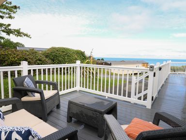 An outdoor deck with black chairs, a small table, cushions, white railing, and a view of mobile homes and the sea at The Potting Shed in New Quay