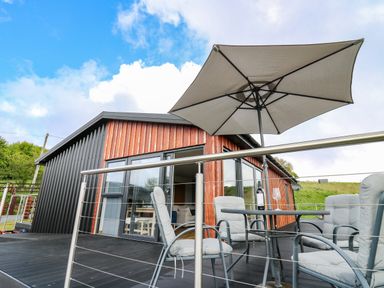 An outdoor patio with cushioned chairs a table with a wine bottle and glass and a large umbrella attached to a modern house at Llyn Cae Ty Nant Fishery Holiday Let Bwlchllan near Llangeitho