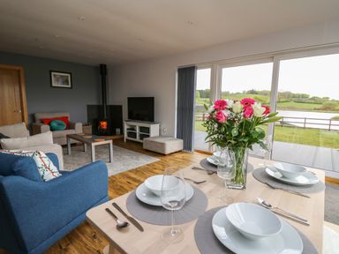 A dining table set with plates bowls glasses and cutlery next to a living room with armchairs a coffee table a wood stove and a television at Llyn Cae Ty Nant Fishery Holiday Let in Bwlchllan near Llangeitho