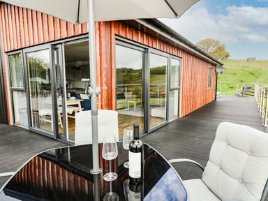 A patio table with chairs and umbrella on a terrace outside a wooden house with sliding glass doors at Llyn Cae Ty Nant Fishery Holiday Let in Bwlchllan near Llangeitho