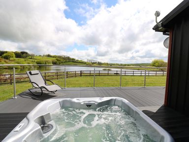 An outdoor deck with a hot tub and a reclining chair overlooking a lake and grassy hills at Llyn Cae Ty Nant Fishery Holiday Let in Bwlchllan near Llangeitho
