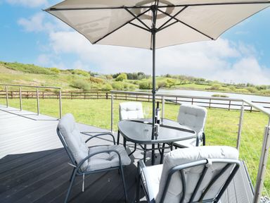 An outdoor patio with a table umbrella four cushioned chairs and a bottle of wine overlooking a grassy area and a lake at Llyn Cae Ty Nant Fishery Holiday Let in Bwlchllan near Llangeitho