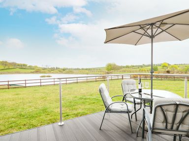 An outdoor patio with cushioned chairs table umbrella and a bottle with glasses overlooking a lake and grassy field at Llyn Cae Ty Nant Fishery Holiday Let in Bwlchllan near Llangeitho