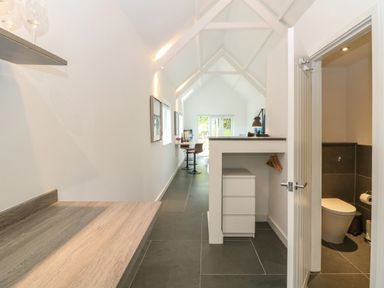 A narrow room with a high vaulted ceiling a desk with drawers a small toilet room and a seating area at Twigs in Woody Bay