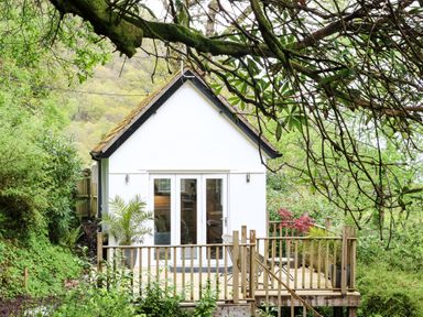 A small white house with a wooden deck surrounded by greenery at Twigs in Woody Bay