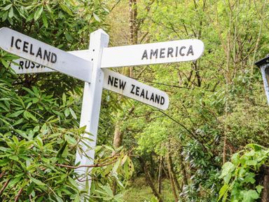 A white wooden signpost with directions to Iceland America New Zealand and Russia surrounded by green trees and bushes