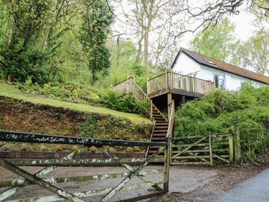 A house with a wooden deck and stairs next to a gated entrance surrounded by trees and bushes at Twigs in Woody Bay
