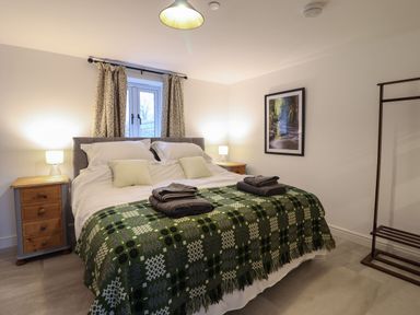 A bedroom with a bed covered by patterned green bedding two bedside tables with lamps a window with curtains and a framed picture on the wall at Merddwr in Cerrigydrudion