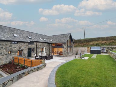 An outdoor patio with table and chairs a stone house and lawn with hot tub and stone wall at Merddwr in Cerrigydrudion