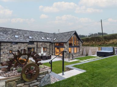 A stone house with large windows a patio with outdoor furniture a hot tub and a grassy yard at Merddwr in Cerrigydrudion