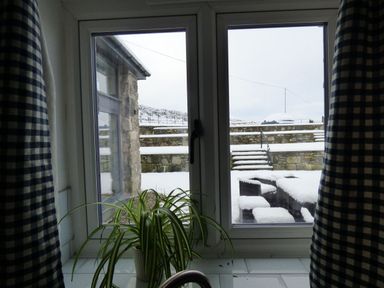 A window with blue checkered curtains looking out to a snowy yard with stone walls and outdoor furniture at Merddwr in Cerrigydrudion