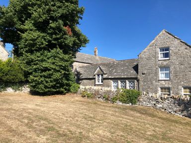 A stone house with multiple windows and a stone wall in front next to a large green tree at Sea Pink Cottage in Langton Matravers