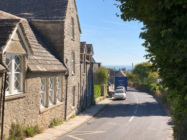 A street with stone houses on the left cars parked on the right and trees on both sides at Sea Pink Cottage in Langton Matravers