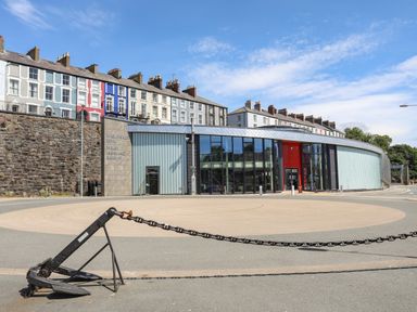A modern building with glass windows and a circular paved area in front along with an anchor and chain in the foreground with houses on a stone wall behind at Welsh Highland Railway in Caernarfon