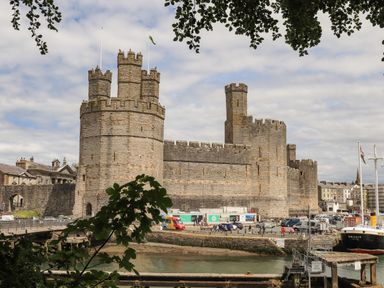 A stone castle with multiple towers near a waterway and parked cars at Nantlle in Caernarfon