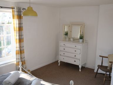 A bedroom corner with a white chest of drawers with a three-panel mirror a wooden chair and a window with striped curtains at Phloxhaven in Reedham